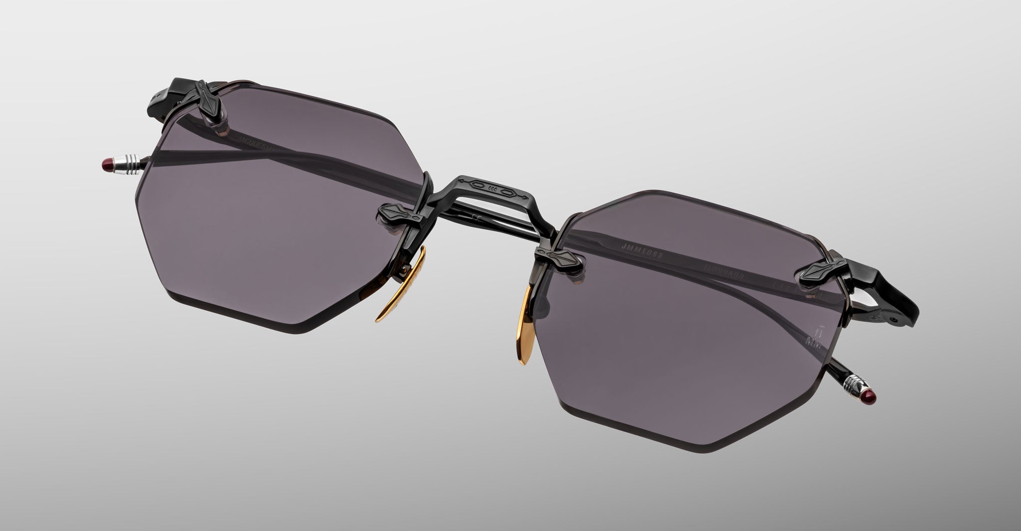 The Jacques Marie Mage El Dorado sunglasses feature geometric hexagonal dark-tinted lenses, thin black metal frames, and gold nose pad accents, showcased on a light gray background.