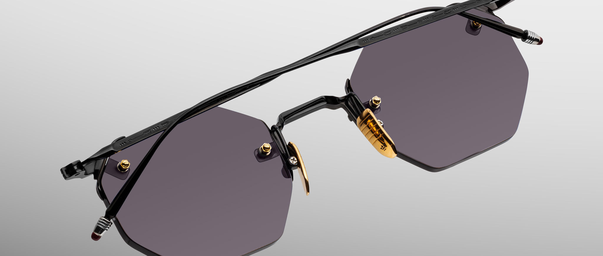 The Jacques Marie Mage El Dorado: sleek sunglasses featuring dark hexagonal lenses, slim black metal frames, gold nose pads, and a minimalist design, displayed on a light gray gradient background.