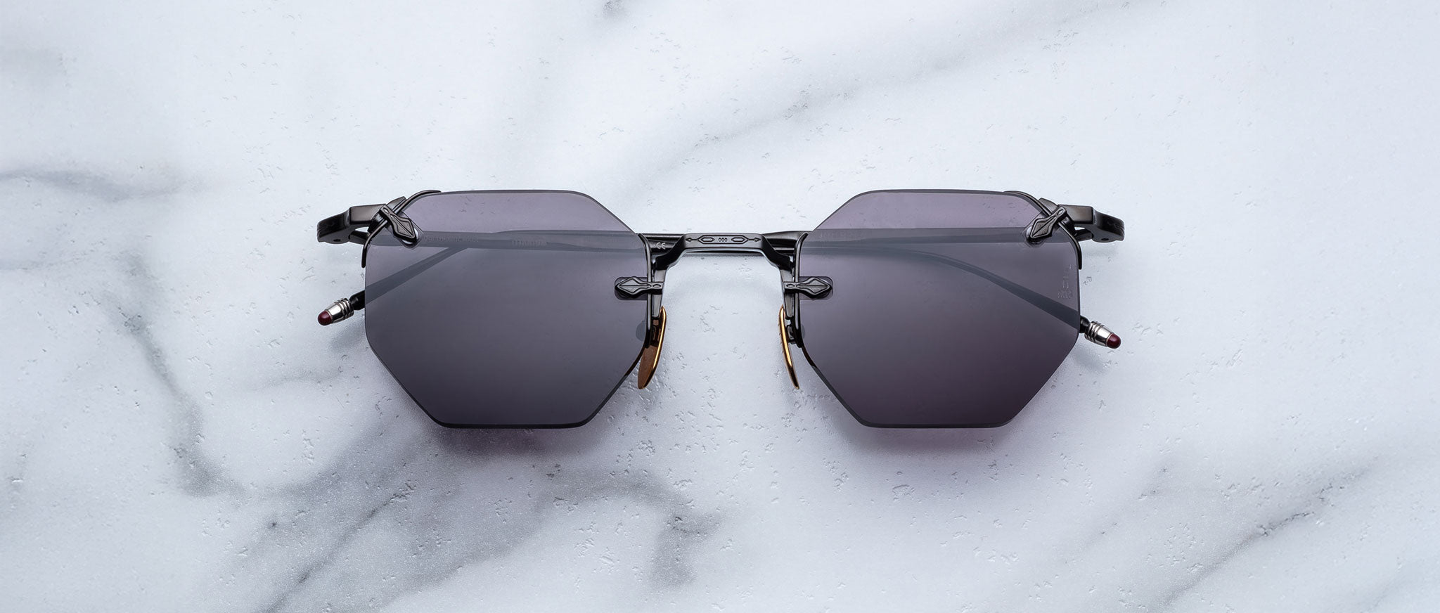 The Jacques Marie Mage El Dorado sunglasses, featuring geometric hexagonal black frames and dark lenses, are displayed on a white marble surface.