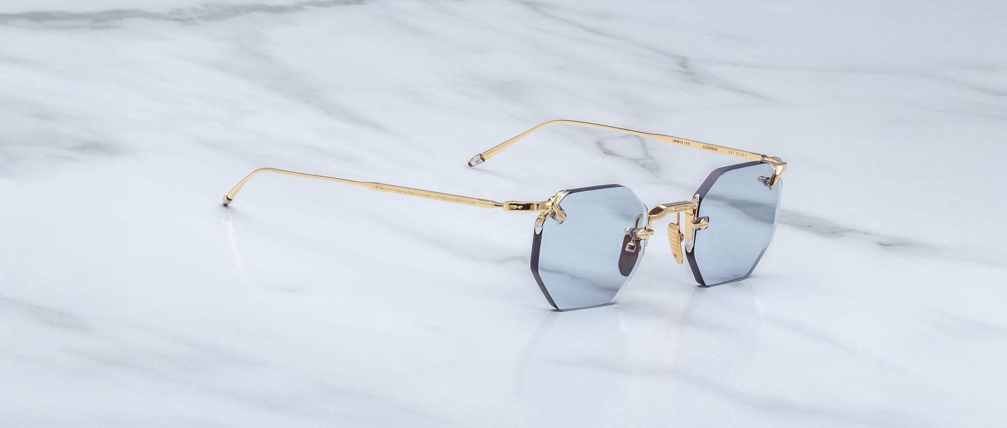 The Jacques Marie Mage El Dorado features octagonal, rimless sunglasses with blue tinted lenses and slim gold metal arms, displayed on a white marble surface.