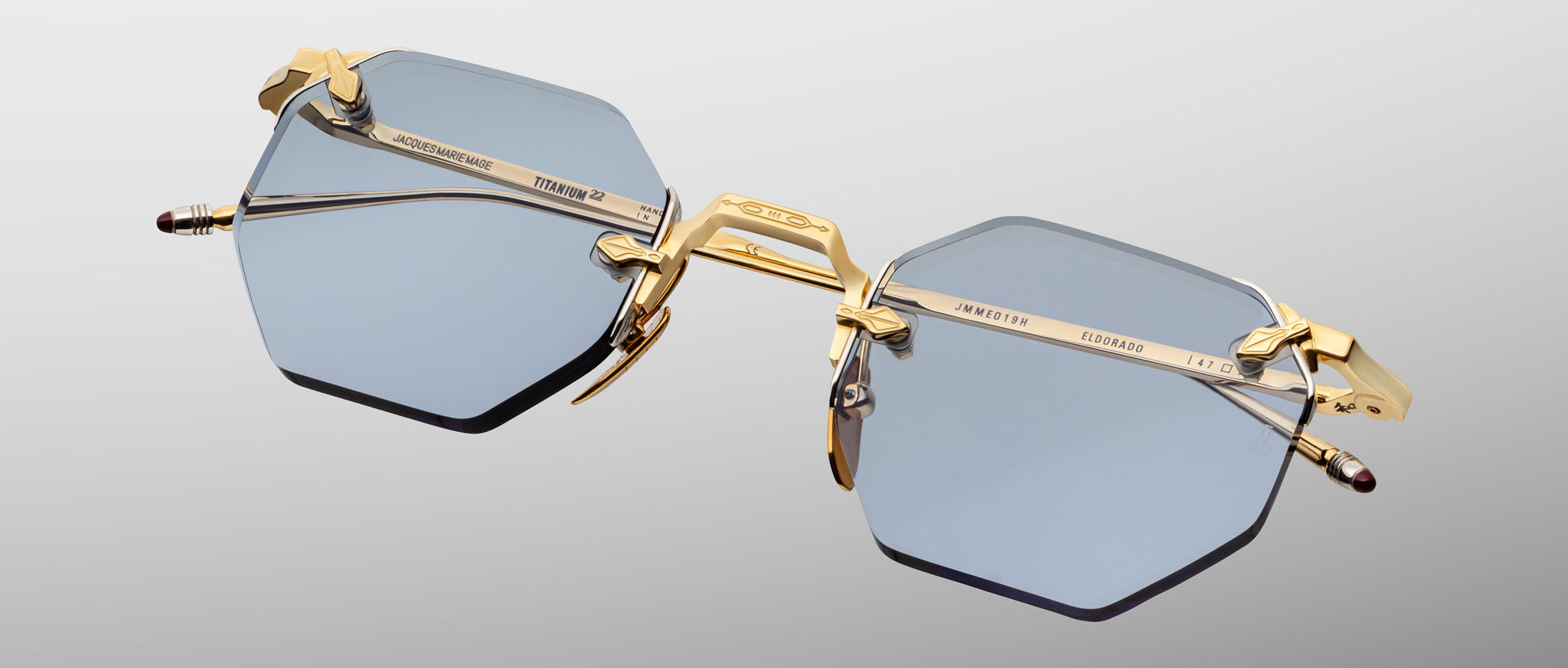 The Jacques Marie Mage El Dorado features rimless hexagonal blue-tinted lenses with gold metal temples and bridge, showcased against a neutral gradient background.