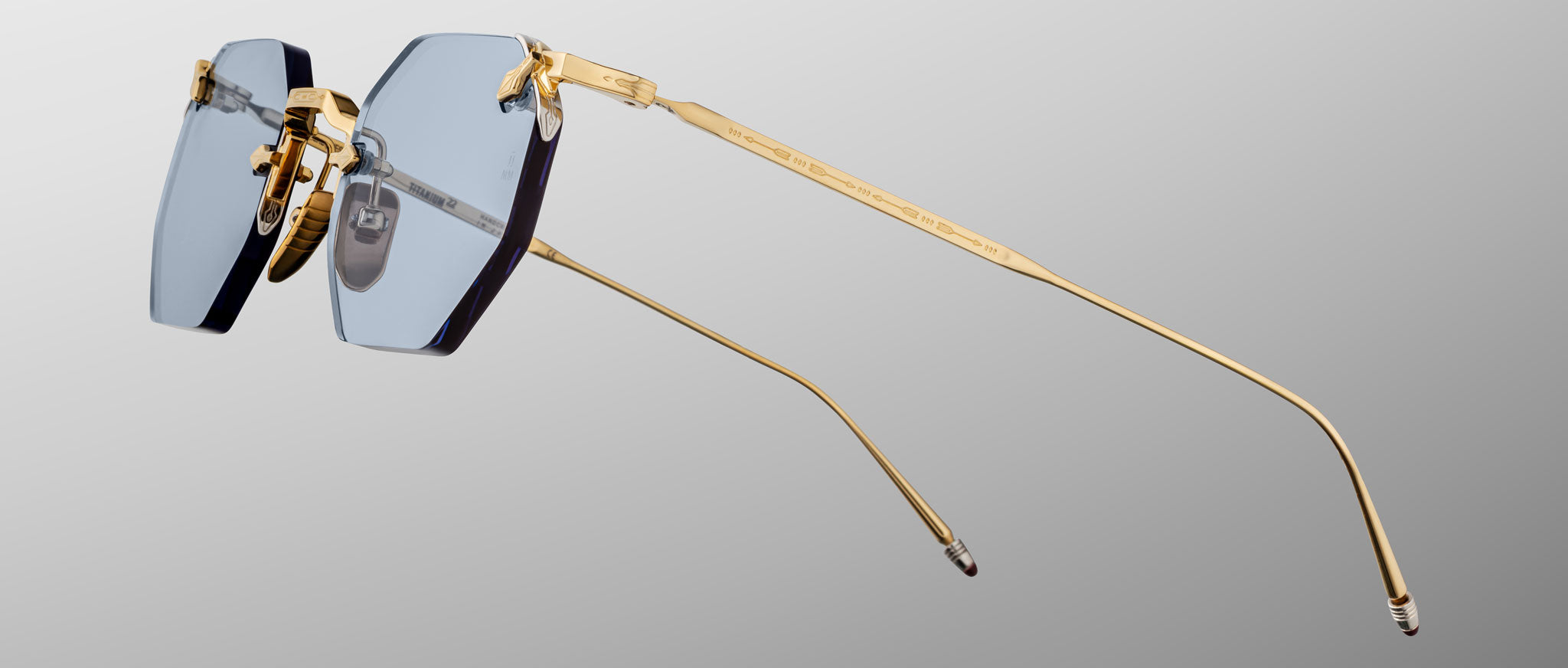 The Jacques Marie Mage El Dorado features rimless eyeglasses with blue-tinted rectangular lenses and slim gold metal arms, displayed against a light gray gradient background.