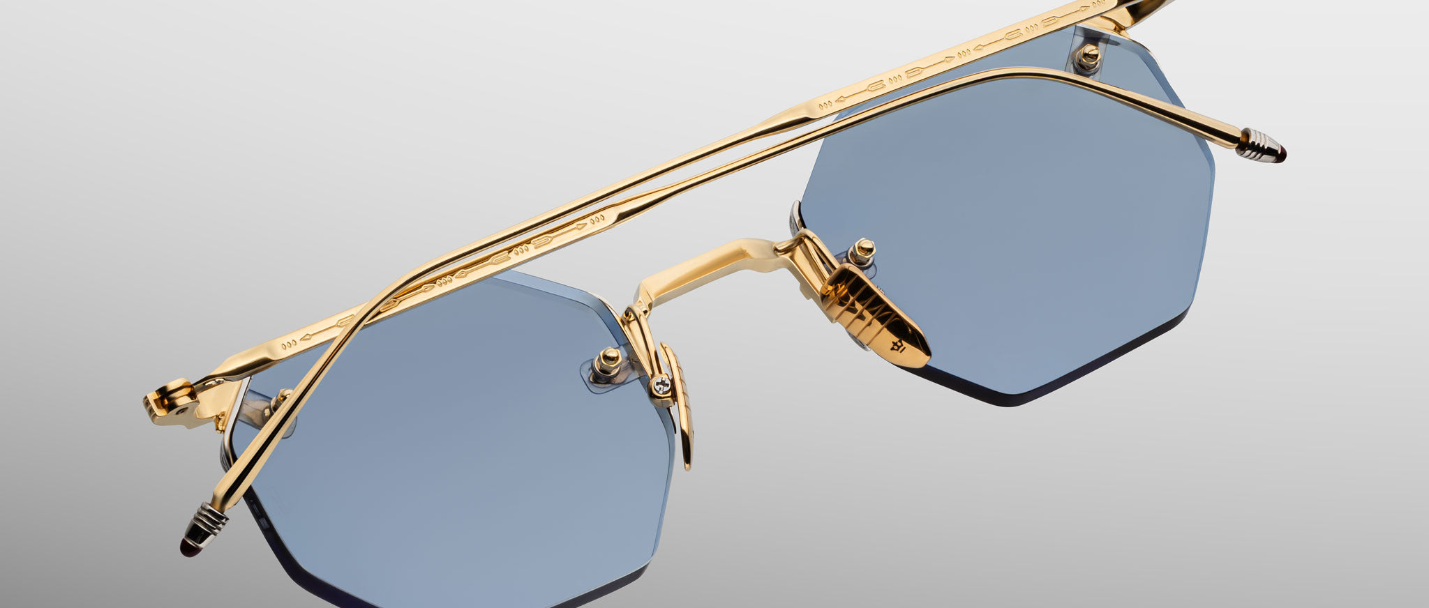 The Jacques Marie Mage El Dorado sunglasses feature a geometric design with blue-tinted lenses and a gold metal frame, showcased from above against a light gradient background.