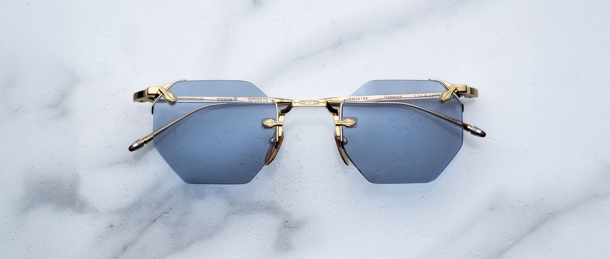 The Jacques Marie Mage El Dorado sunglasses, featuring hexagonal blue lenses and gold metal frames, are displayed on a white marble surface.