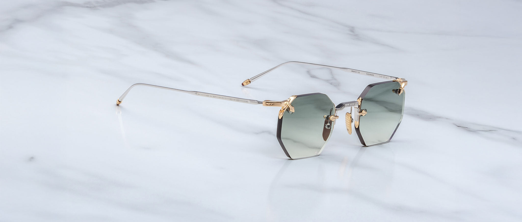 A pair of Jacques Marie Mage El Dorado geometric rimless sunglasses with gray gradient lenses and thin gold-toned arms rests on a white marble surface.