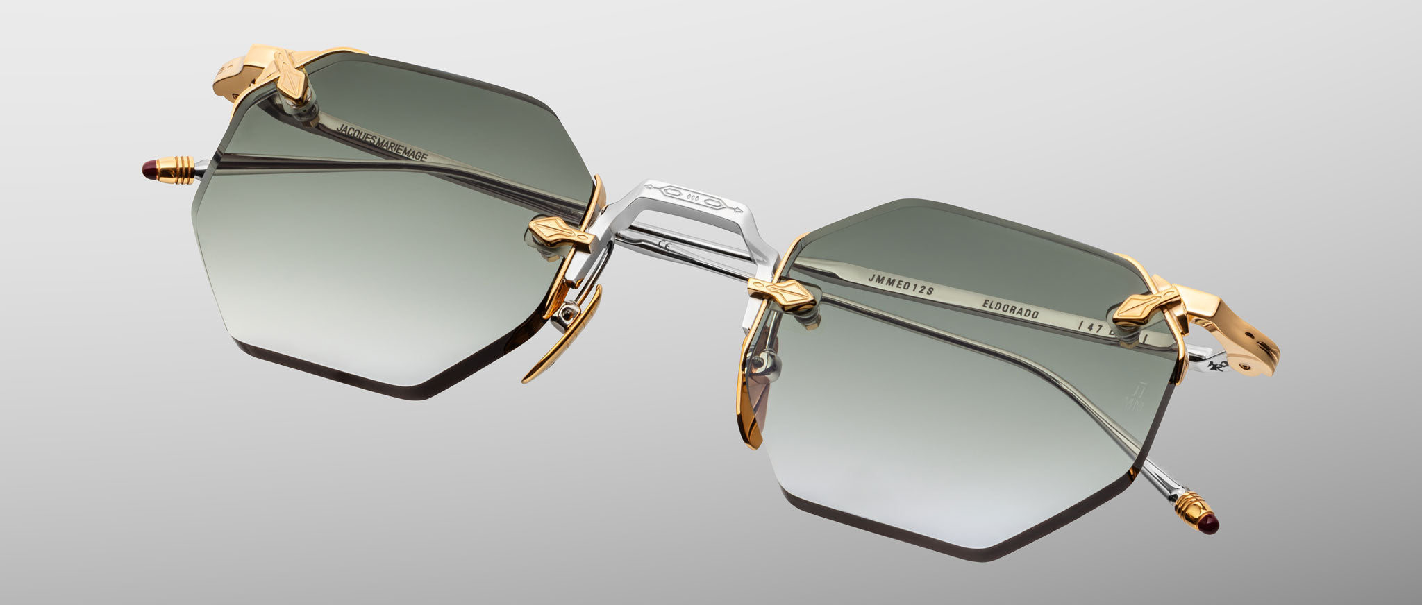 The Jacques Marie Mage El Dorado features hexagonal sunglasses with gradient gray lenses, gold and silver metal frames, and gold accents on the hinges and nose pads, shown against a light gray background.