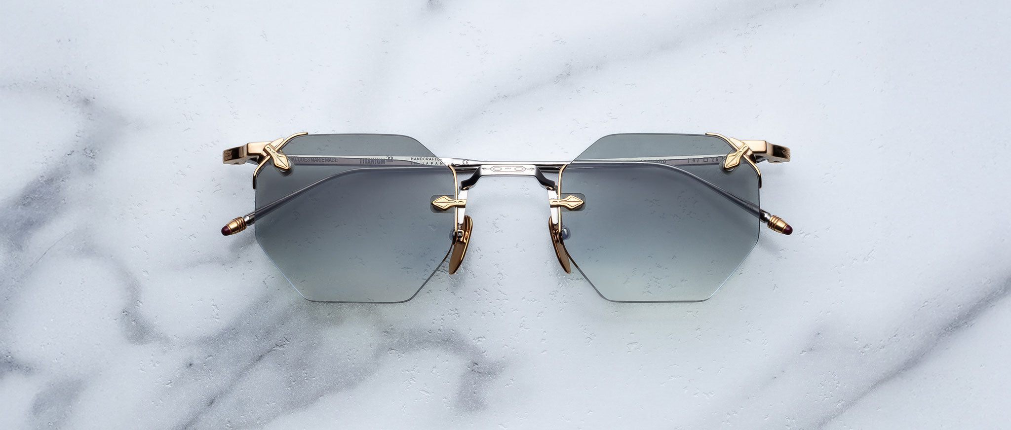 The Jacques Marie Mage El Dorado sunglasses, featuring geometric hexagonal frames with gray lenses and gold accents, are displayed on a white marble surface.