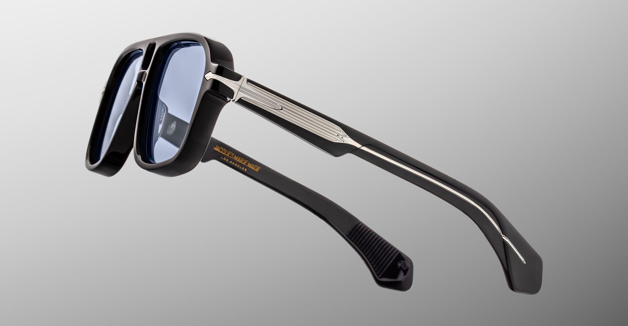 Side view of the Jacques Marie Mage Elkhart limited-edition navigator sunglasses, featuring blue-tinted lenses and a sculptural brow ridge, set against a gray gradient background.