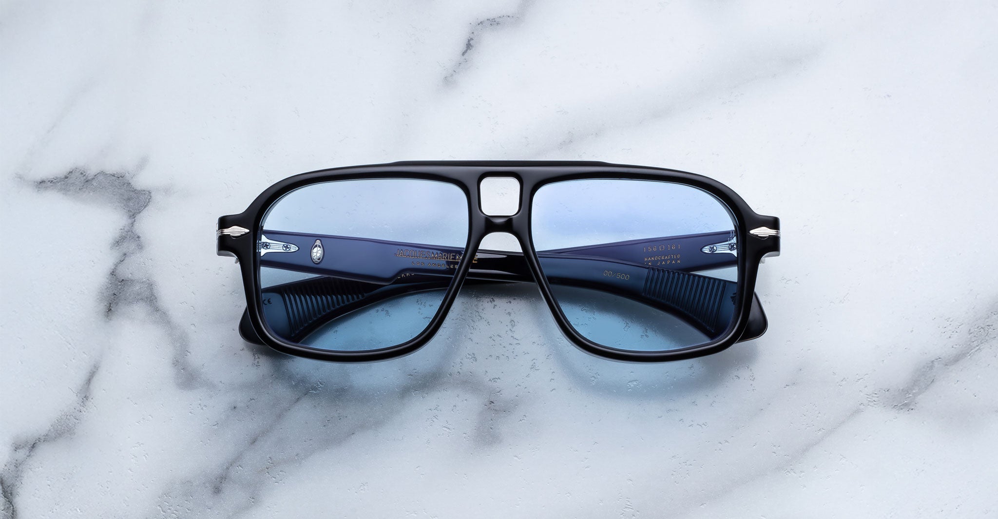 A pair of Jacques Marie Mage Elkhart limited-edition navigator sunglasses with black frames, sculptural brow ridge, and blue-tinted lenses rests on a subtly veined white marble surface.