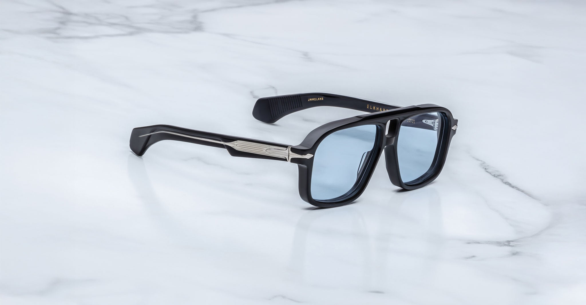 The Jacques Marie Mage Elkhart sunglasses, featuring a black rectangular navigator design with a sculptural brow ridge, blue-tinted lenses, and silver arm accents, are displayed elegantly on a white marble surface.