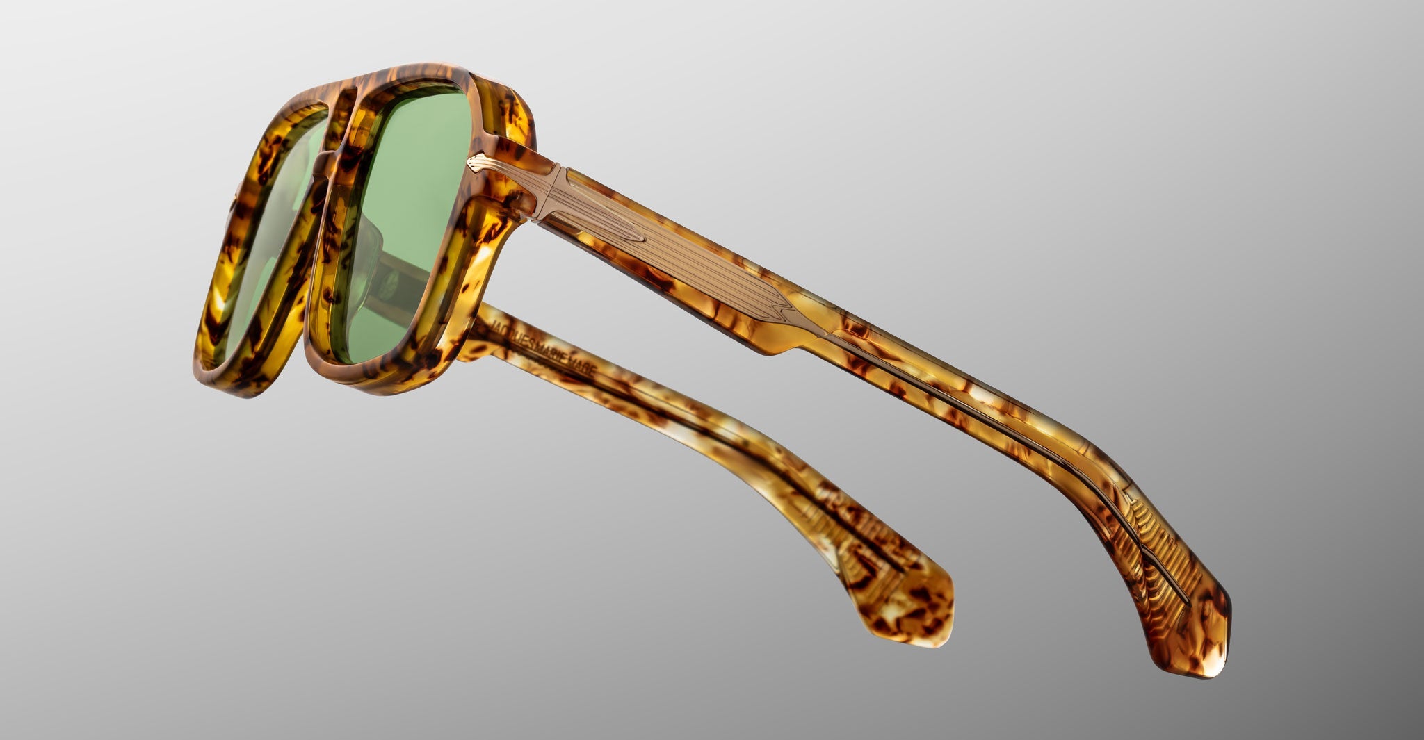 The Jacques Marie Mage Elkhart limited-edition navigator sunglasses feature a tortoiseshell frame, sculptural brow ridge, and green lenses, displayed from an angled side view against a gray background.