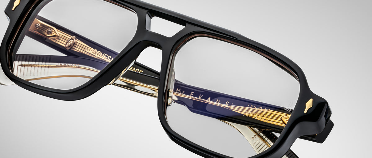 Close-up of Jacques Marie Mage Evans handcrafted optical glasses with an oversized acetate frame and gold and dark blue temple accents, set against a plain light background.