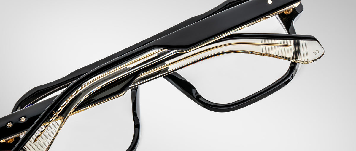 Two pairs of modern eyeglasses, including the Jacques Marie Mage Evans oversized acetate frame in black and gold, are stacked together on a plain gray background.