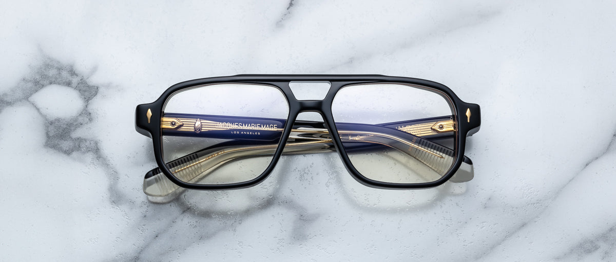 Jacques Marie Mage Evans: Oversized black-rimmed handcrafted optical glasses with gold and navy blue acetate arms by Jacques Marie Mage, displayed on a white marble surface.
