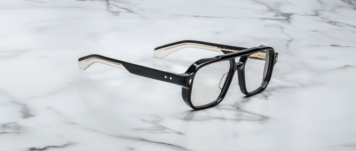 The Jacques Marie Mage Evans optical glasses feature an oversized black and gold acetate frame with clear lenses, showcased on a white marble surface.
