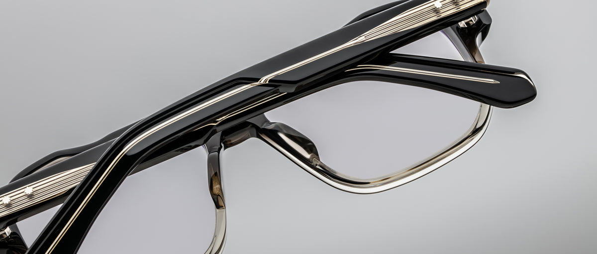 Close-up of Jacques Marie Mage Evans optical glasses, showcasing a black and gold oversized acetate frame with geometric lines and clear lenses, set against a plain gray background.