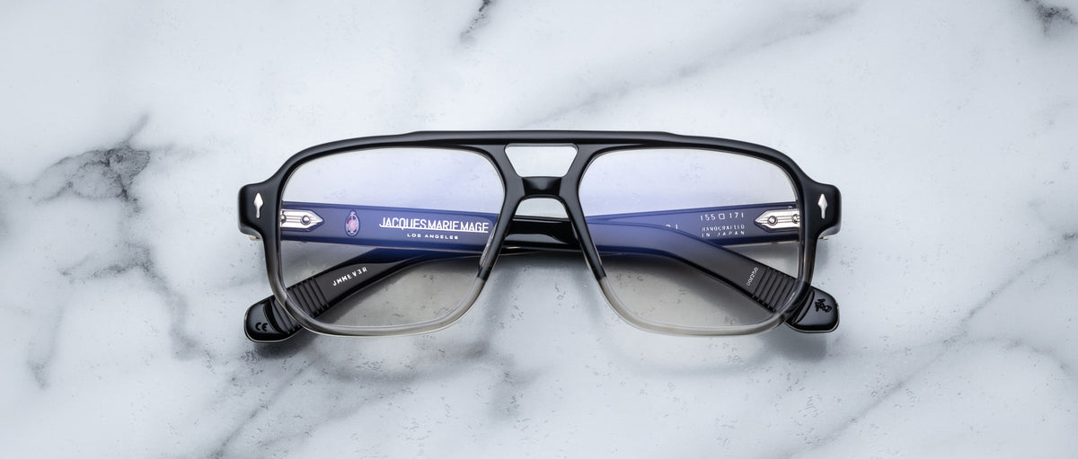 A pair of Jacques Marie Mage Evans eyeglasses rests on white marble, featuring oversized handcrafted acetate frames with the Jacques Marie Mage branding visible inside.
