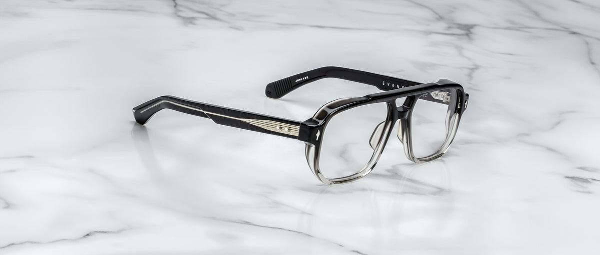 The Jacques Marie Mage Evans eyeglasses by Jacques Marie Mage feature a black and clear ombré oversized acetate frame with thick rectangular silhouette and silver temple detailing, handcrafted and displayed on a white marble surface.