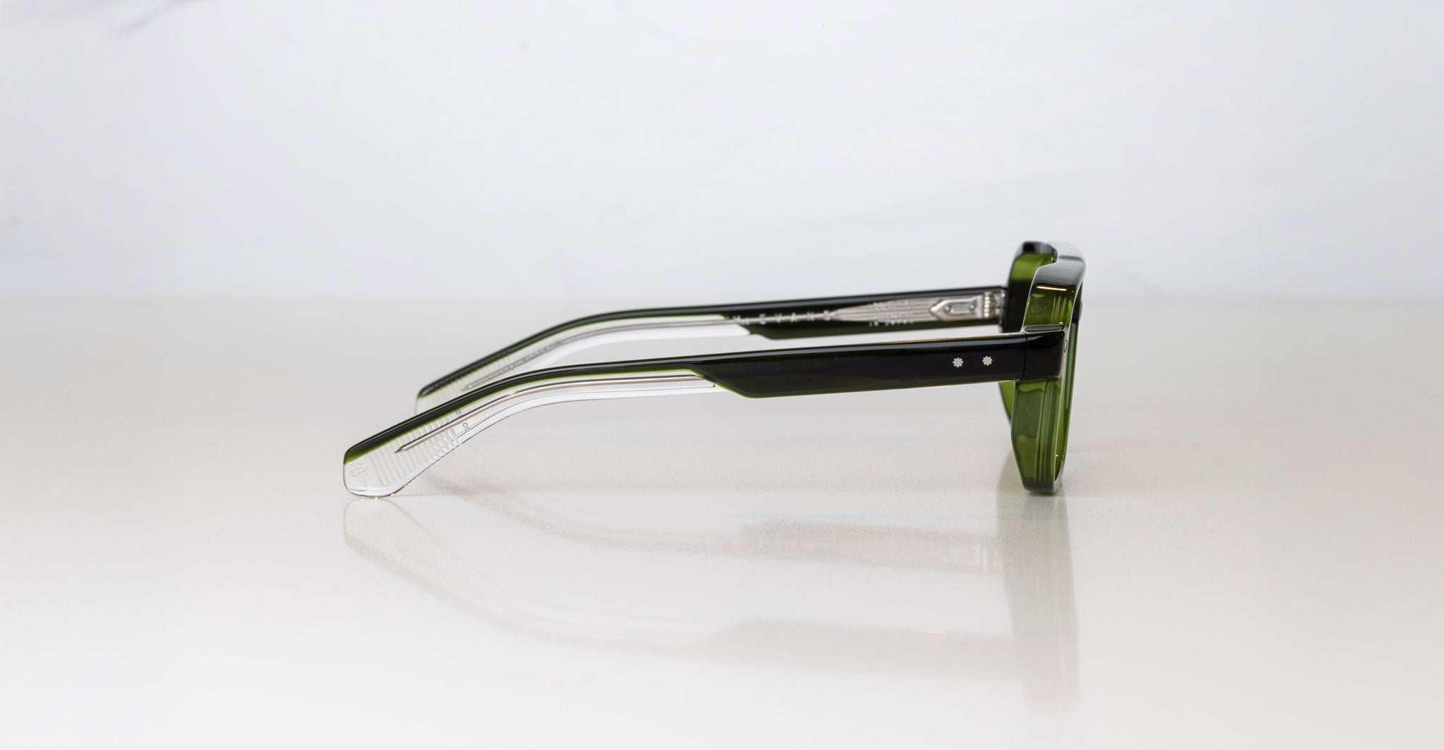 A pair of Jacques Marie Mage Evans green-framed handcrafted optical glasses rests on its side against a plain white background.