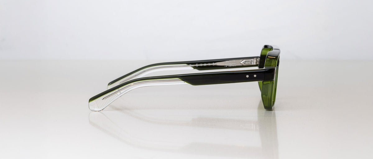 A pair of Jacques Marie Mage Evans green-framed handcrafted optical glasses rests on its side against a plain white background.