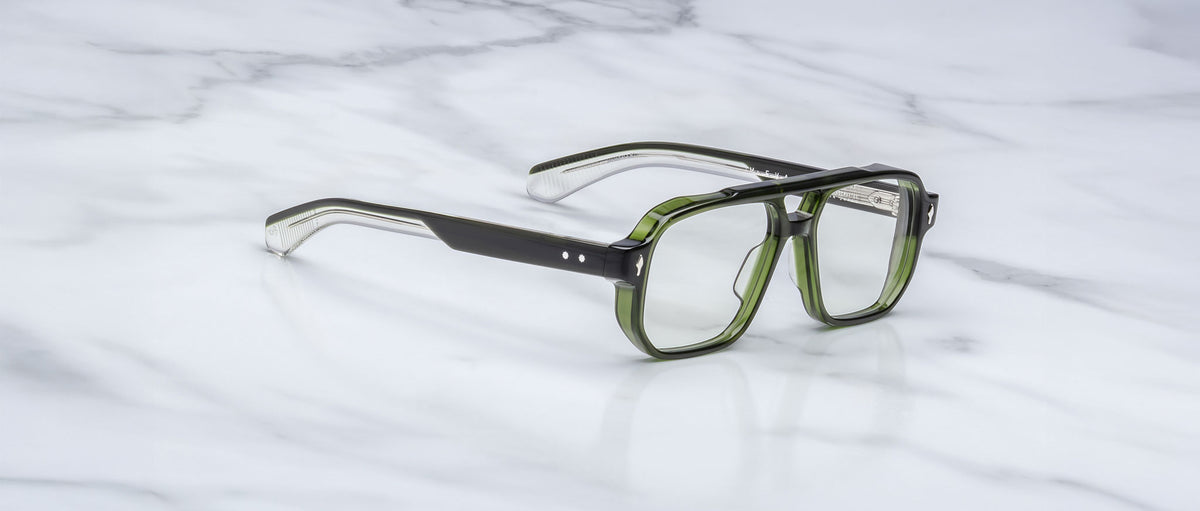 A pair of Jacques Marie Mage Evans handcrafted optical glasses in green and black with thick frames rests on a white marble surface.