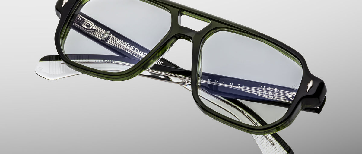 The Jacques Marie Mage Evans eyeglasses feature an oversized rectangular acetate frame with green rims and black-and-white patterned arms, shown against a grey background.