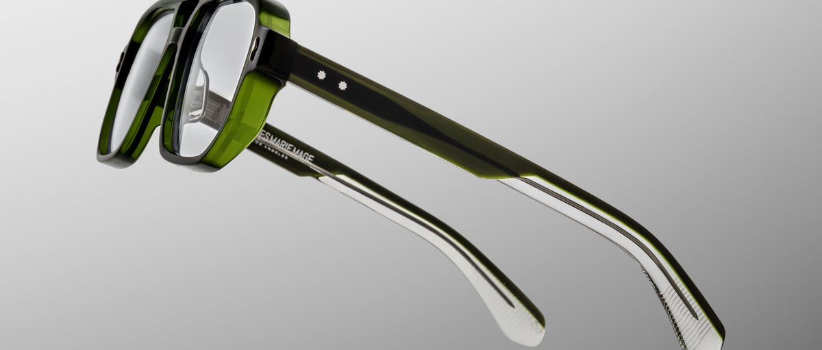 The Jacques Marie Mage Evans optical glasses feature handcrafted green and clear acetate rectangular frames, displayed against a gray gradient background.