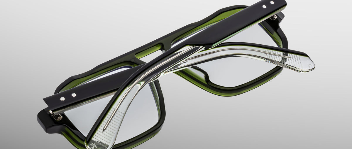 The Jacques Marie Mage Evans optical glasses feature handcrafted thick black and green frames, shown folded with crossed arms against a neutral gradient background.