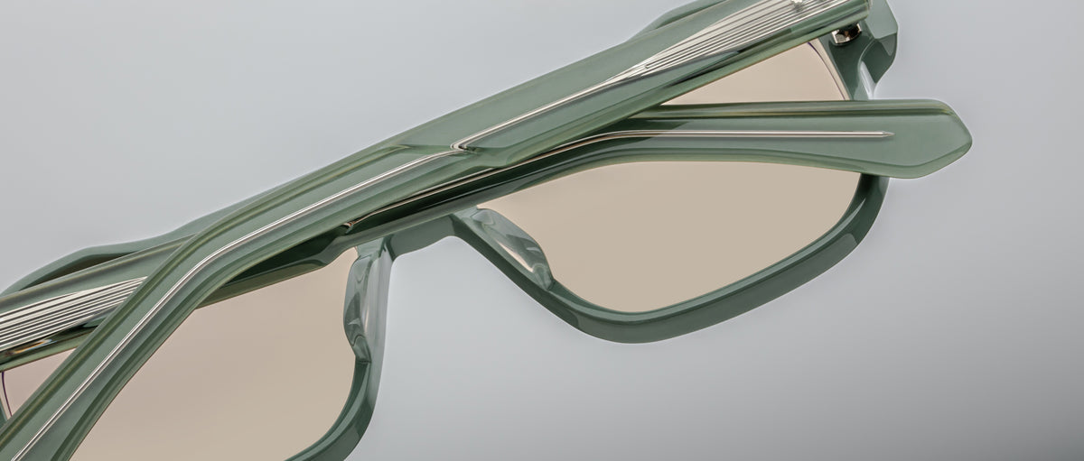 Close-up of Jacques Marie Mage Evans sunglasses: modern, light green acetate frames with beige lenses and overlapping hinges, shown against a neutral background and reflecting the distinctive Jacques Marie Mage style.