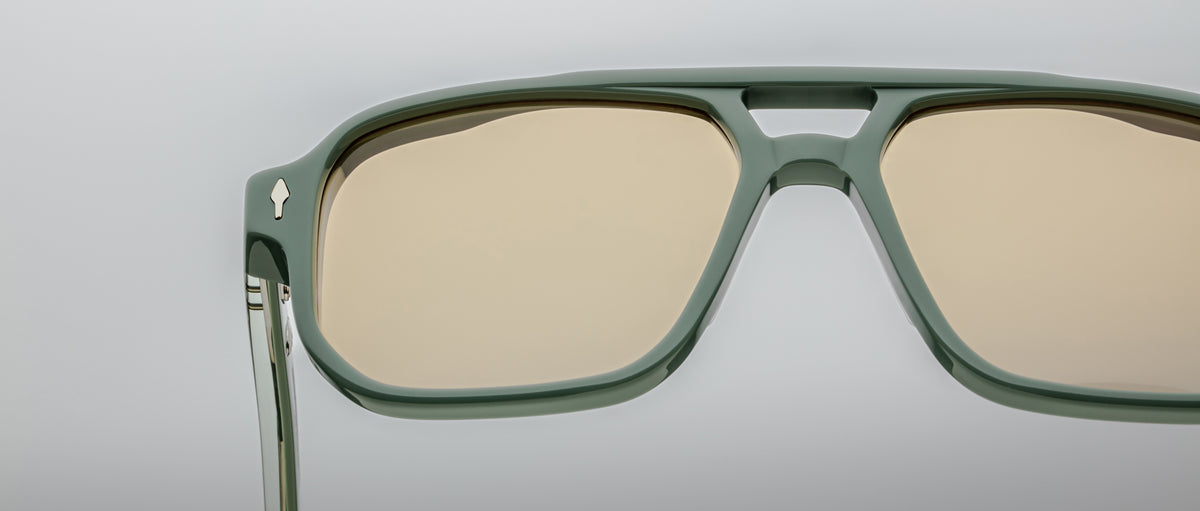 Close-up of Jacques Marie Mage Evans sunglasses with oversized green acetate frames and beige-tinted lenses, set against a plain light gray background.