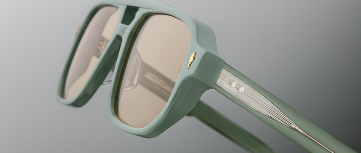 Close-up of Jacques Marie Mage Evans sunglasses in mint green with an oversized acetate frame, gold accents, and light brown lenses set against a gradient gray background.