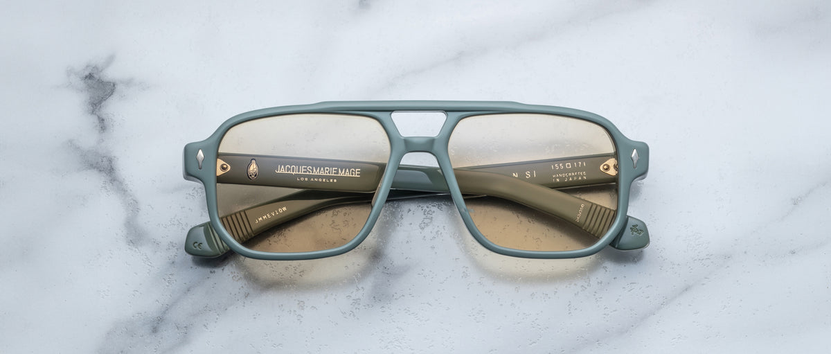 The Jacques Marie Mage Evans eyeglasses by Jacques Marie Mage feature an oversized matte green acetate rectangular frame with gold accents, displayed on a white marble surface.