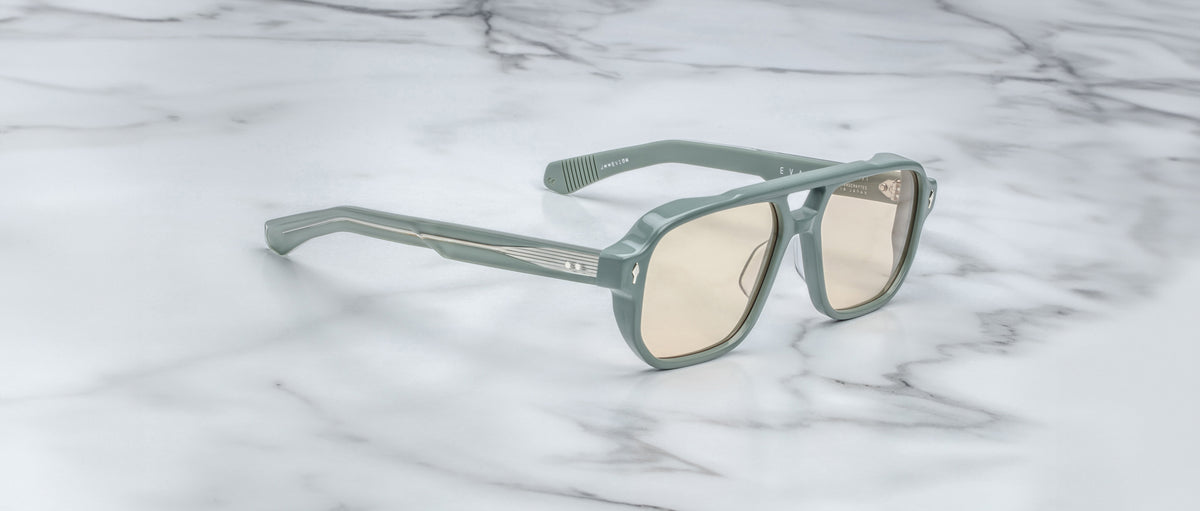 The Jacques Marie Mage Evans sunglasses, featuring an oversized light green acetate frame and beige-tinted lenses, rest on a white marble surface.