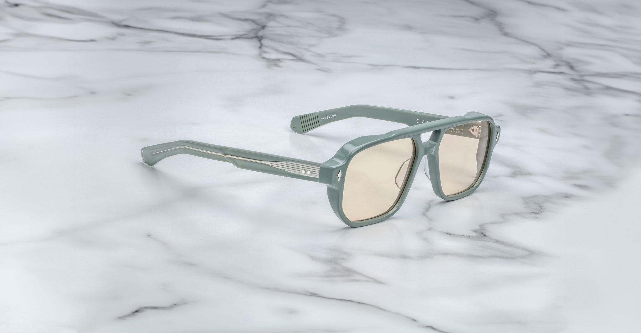 The Jacques Marie Mage Evans sunglasses, featuring an oversized light green acetate frame and beige-tinted lenses, rest on a white marble surface.