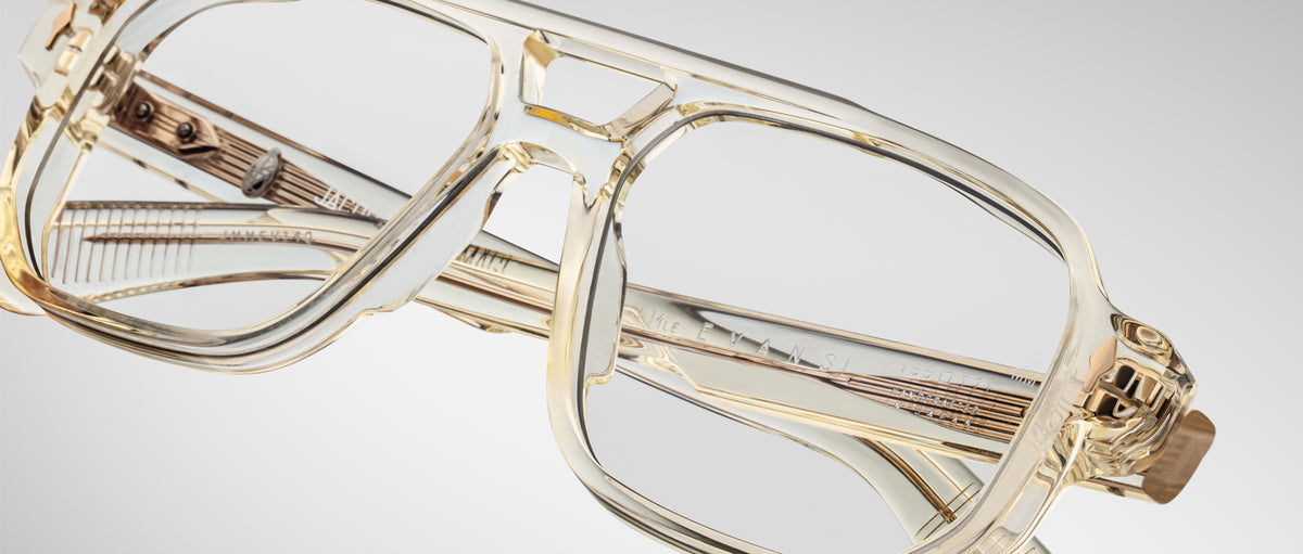 The Jacques Marie Mage Evans eyeglasses feature a clear, oversized acetate frame with thick, transparent rims and large lenses, showcased against a plain, light gray background.