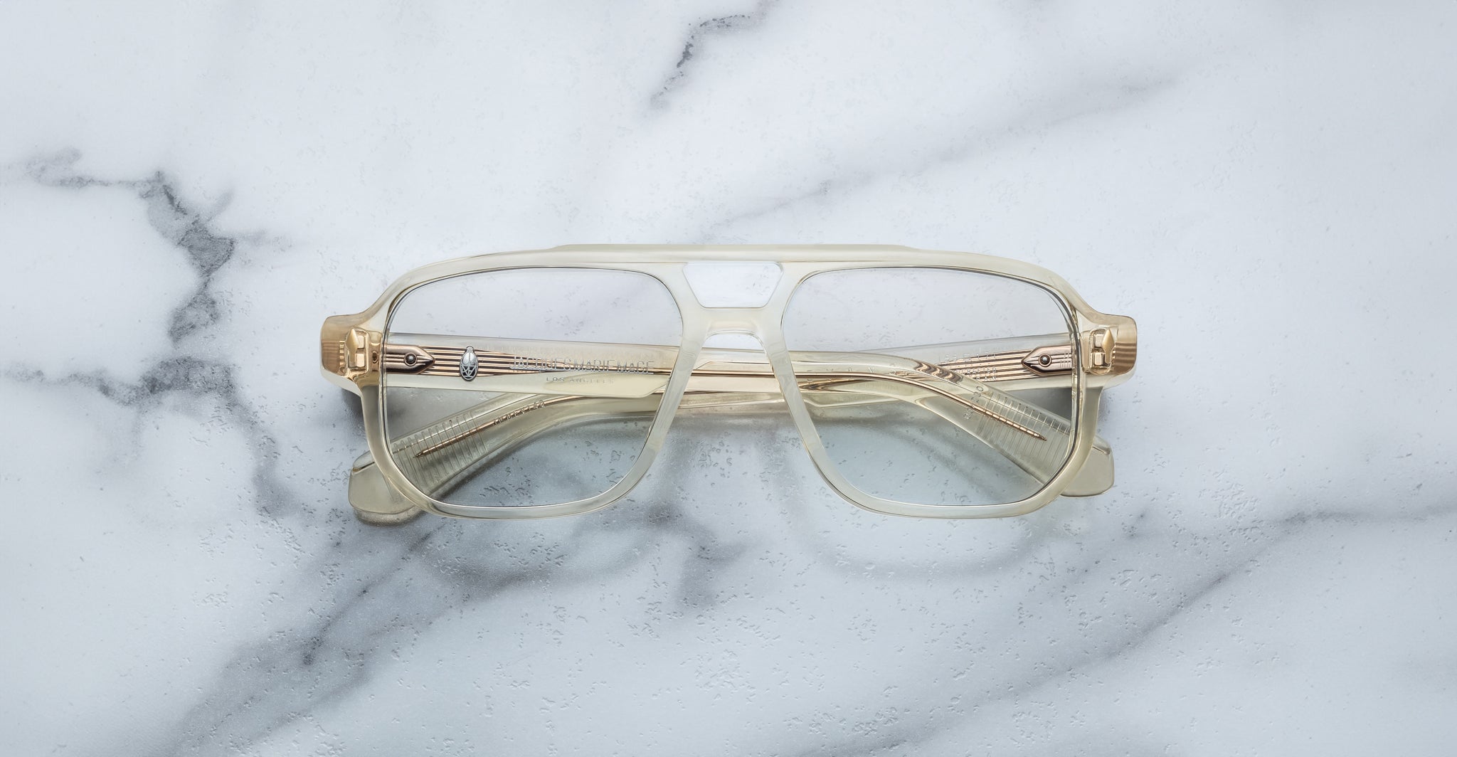The Jacques Marie Mage Evans are handcrafted optical glasses with clear oversized acetate frames and gold accents, elegantly displayed on a white marble surface.
