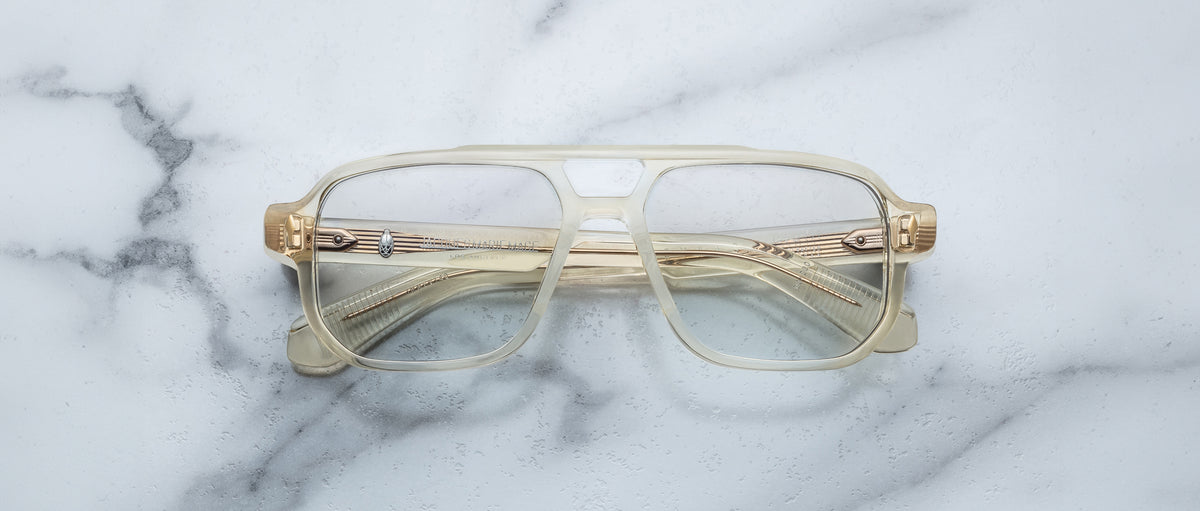 The Jacques Marie Mage Evans are handcrafted optical glasses with clear oversized acetate frames and gold accents, elegantly displayed on a white marble surface.