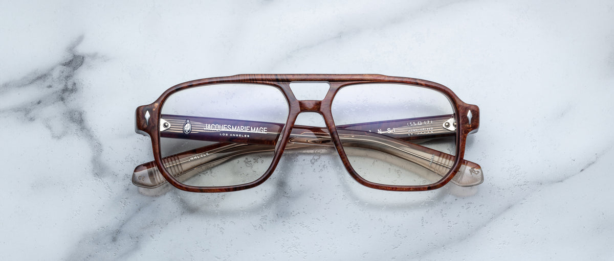 Jacques Marie Mage Evans handcrafted optical glasses with oversized brown acetate frames and clear lenses rest on a white marble surface, their folded arms neatly placed behind the frames.