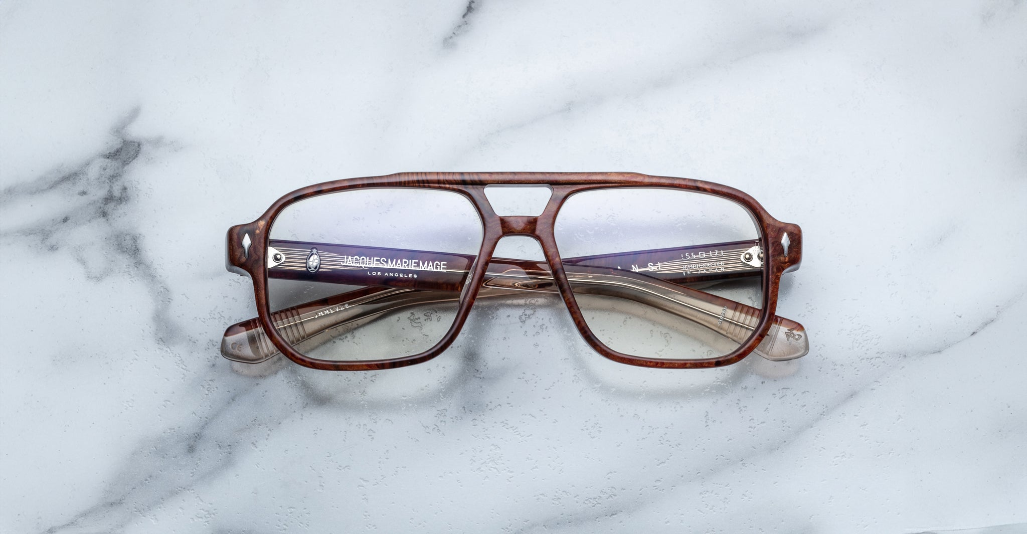 Jacques Marie Mage Evans handcrafted optical glasses with oversized brown acetate frames and clear lenses rest on a white marble surface, their folded arms neatly placed behind the frames.