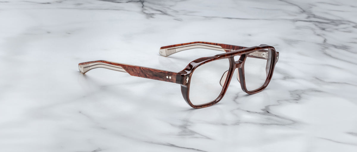The Jacques Marie Mage Evans eyeglasses by Jacques Marie Mage feature an oversized brown acetate frame and clear lenses, displayed on a white marble surface to highlight their handcrafted optical design.