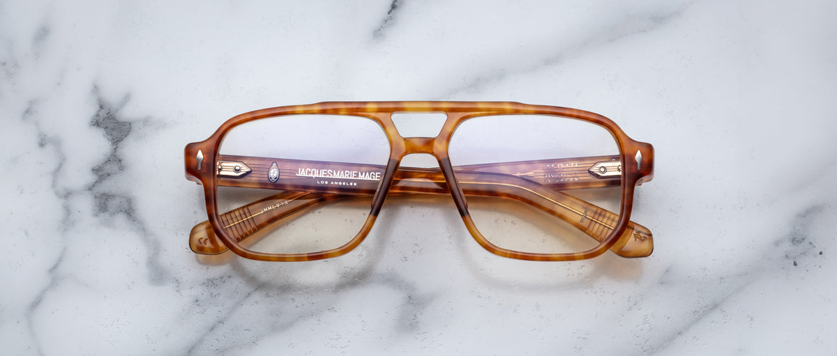 The Jacques Marie Mage Evans eyeglasses feature thick, oversized brown tortoiseshell rectangular acetate frames and are shown on a white marble surface.