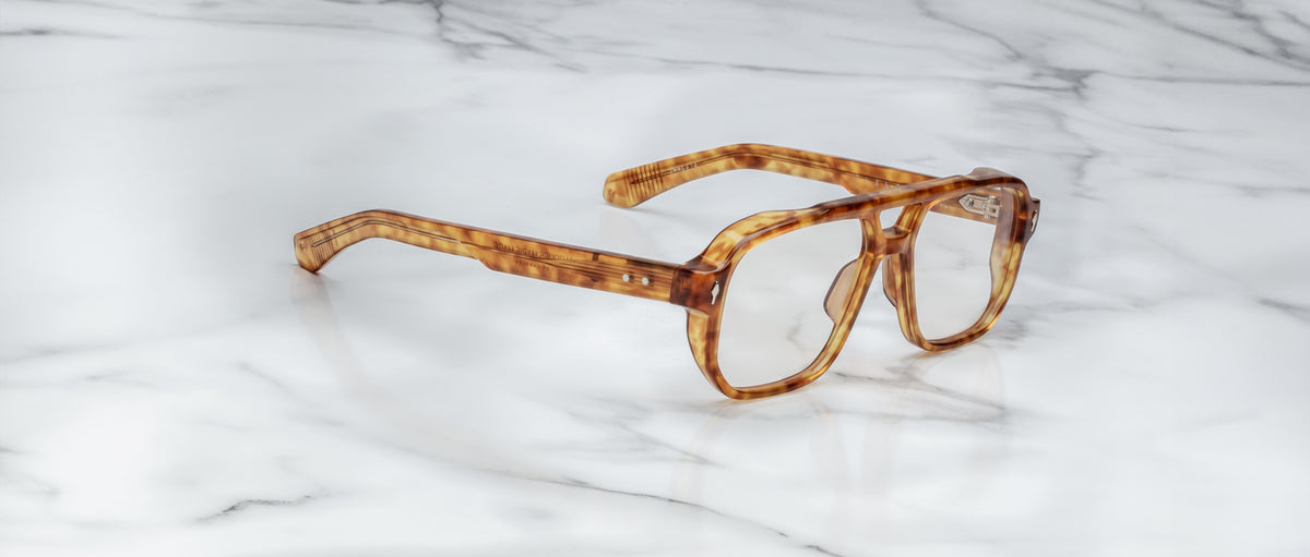 Jacques Marie Mage Evans oversized acetate eyeglasses in brown tortoiseshell by Jacques Marie Mage are displayed on a white marble surface, highlighting their handcrafted optical style.