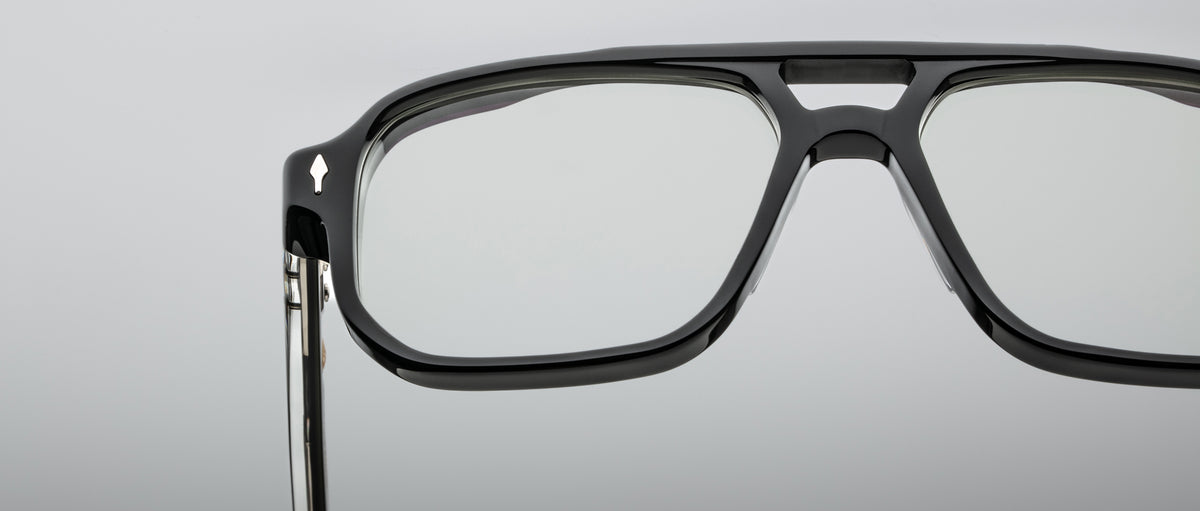 Close-up of the upper half of Jacques Marie Mage Evans, black thick-rimmed oversized acetate eyeglasses with clear lenses, set against a plain gray background.