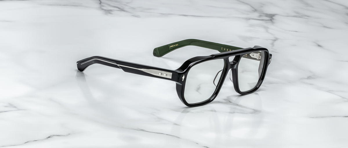 The Jacques Marie Mage Evans glasses feature handcrafted rectangular lenses, silver temple accents, and green inner arms, showcased on a white marble surface.