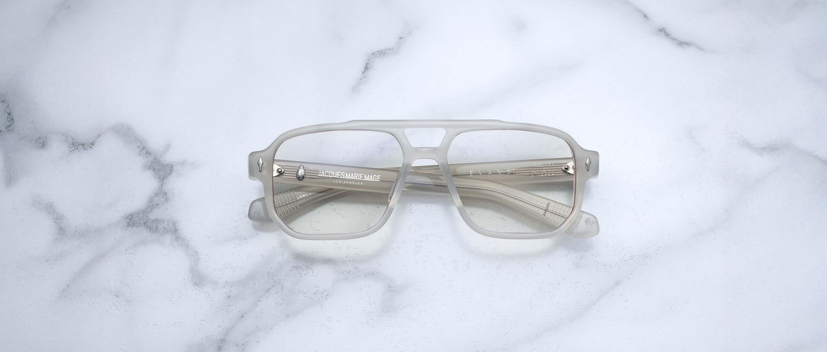 The Jacques Marie Mage Evans eyeglasses by Jacques Marie Mage feature oversized, translucent square acetate frames and are displayed on a white marble surface.