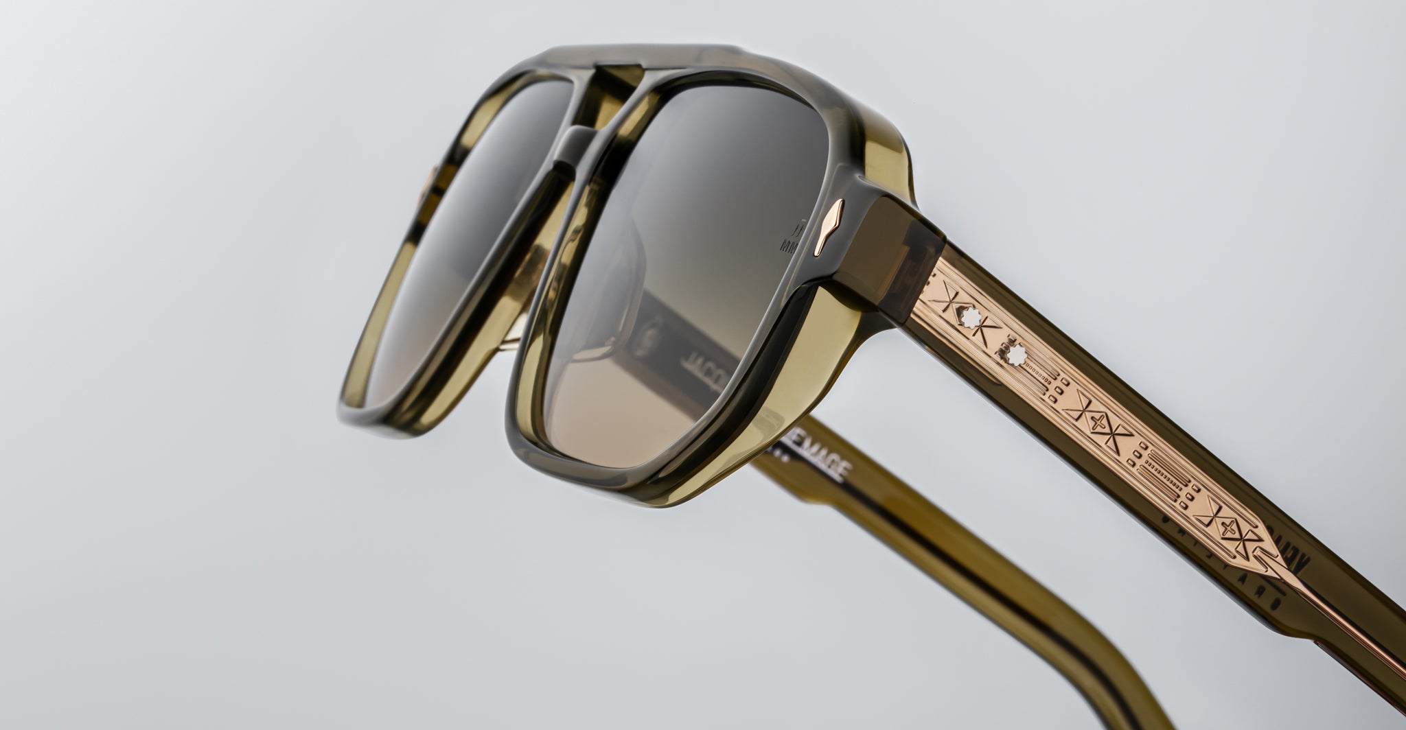 Close-up of Jacques Marie Mage Evans sunglasses in olive green with an oversized acetate frame, dark lenses, and gold-detailed temples engraved with symbols, photographed against a plain background.
