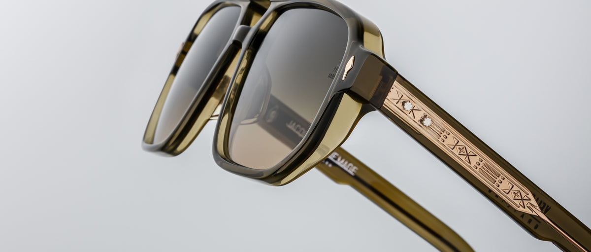 Close-up of Jacques Marie Mage Evans sunglasses in olive green with an oversized acetate frame, dark lenses, and gold-detailed temples engraved with symbols, photographed against a plain background.