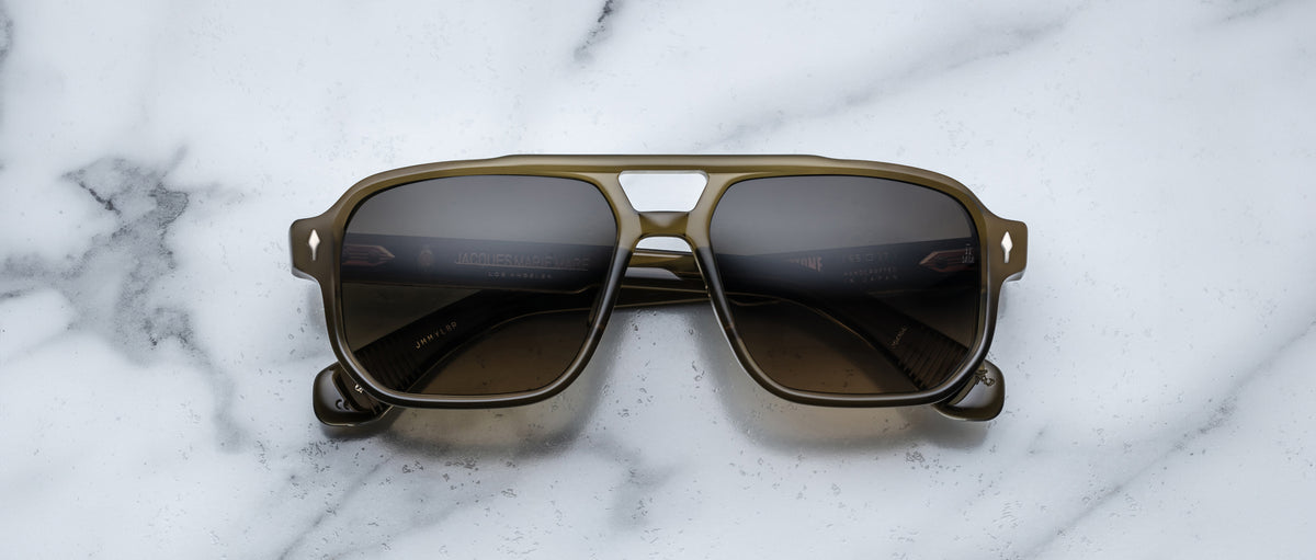 A pair of Jacques Marie Mage Evans sunglasses with dark lenses, an oversized acetate frame, and a greenish tint rests on a white marble surface.