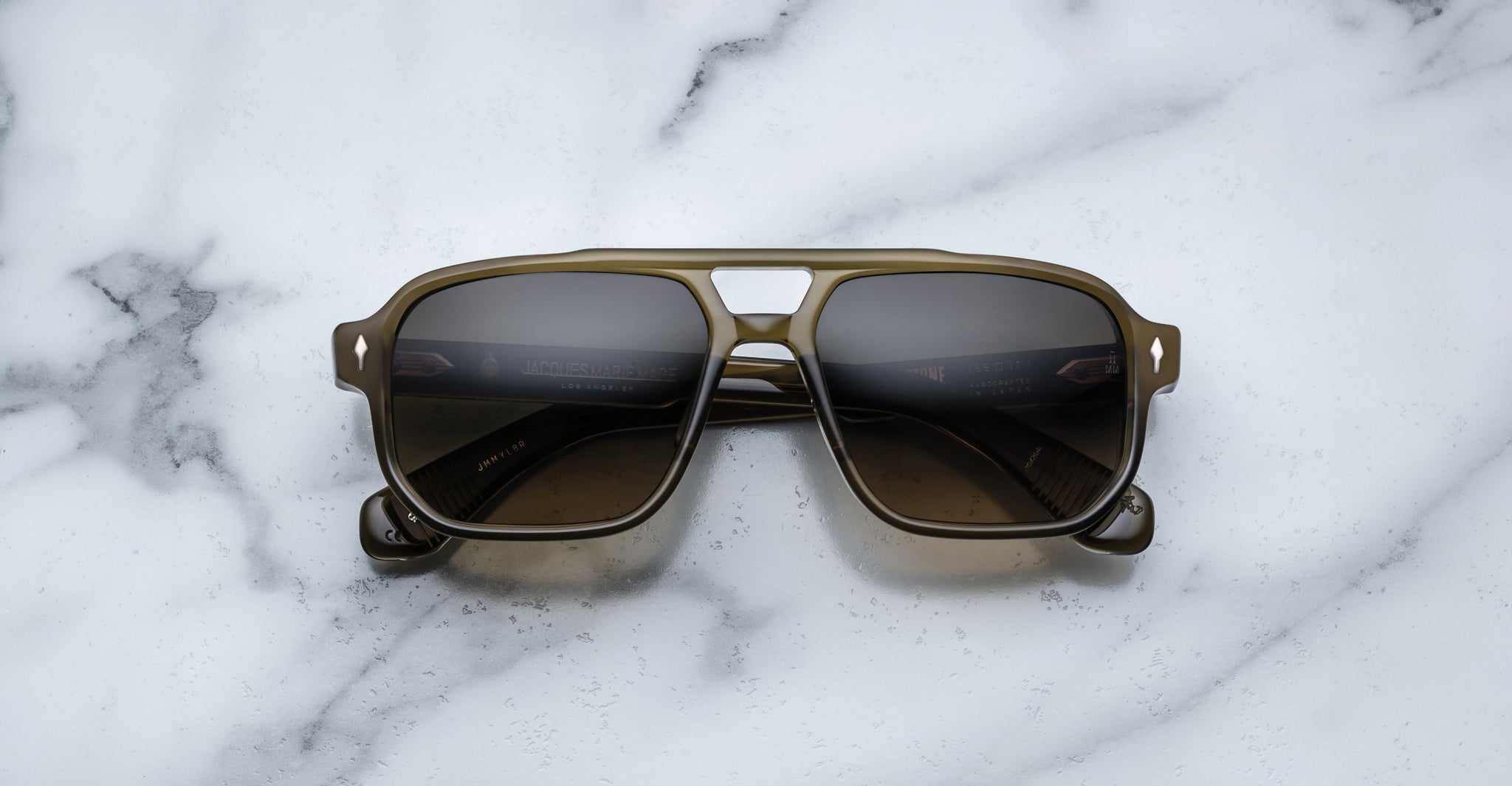 A pair of Jacques Marie Mage Evans sunglasses with dark lenses, an oversized acetate frame, and a greenish tint rests on a white marble surface.