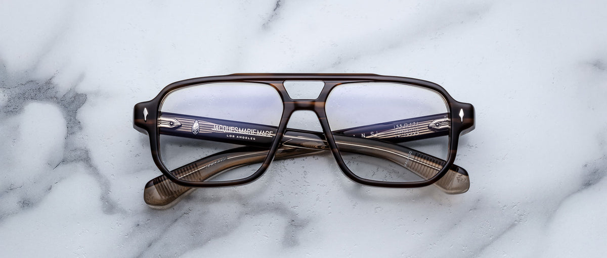 The Jacques Marie Mage Evans eyeglasses by Jacques Marie Mage feature a brown, oversized rectangular acetate frame, displayed on a white marble surface.
